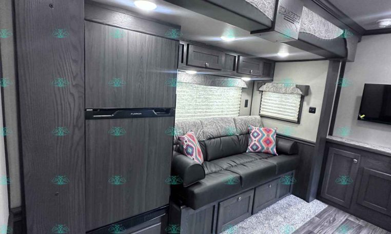 Modern RV interior with dark wood cabinetry, gray sofa with colorful patterned pillows, and overhead storage; built-in TV area and kitchen cabinets run along the wall.