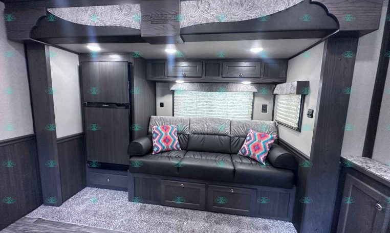 Dark wood RV living area with a black leather sofa, patterned cushions, and overhead cabinets under a corrugated ceiling and recessed lighting.