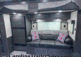 RV living area with dark wood cabinets, gray sofa and colorful geometric pillows, window with blinds, and overhead storage cabinets.