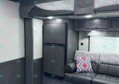 Interior of a dark-wood RV with a built-in refrigerator, overhead cabinets, decorative paneling that reads 'Stampede by Merhow', and a gray sofa with a colorful patterned pillow.
