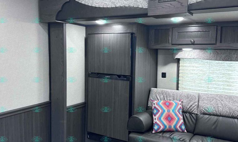 Interior of a dark-wood RV with a built-in refrigerator, overhead cabinets, decorative paneling that reads 'Stampede by Merhow', and a gray sofa with a colorful patterned pillow.