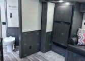 Interior of a modern travel trailer: small bathroom with toilet on left, dark wood cabinets, and a compact fridge on the right; seating with pillows nearby.