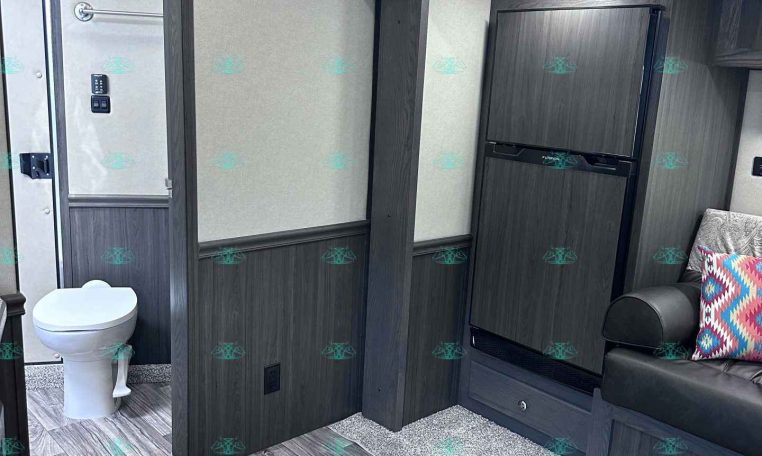 Interior of a modern travel trailer: small bathroom with toilet on left, dark wood cabinets, and a compact fridge on the right; seating with pillows nearby.