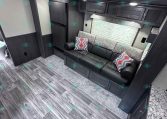 Interior of a dark-wood RV living area with a gray patterned sofa, colorful throw pillows, and overhead cabinets.