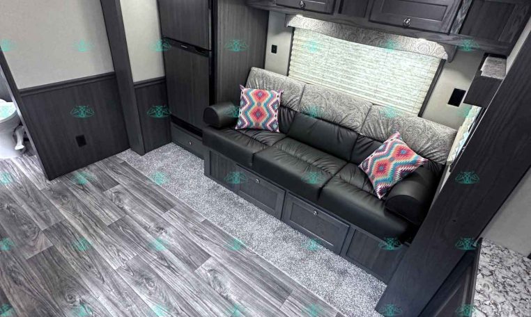 Interior of a dark-wood RV living area with a gray patterned sofa, colorful throw pillows, and overhead cabinets.