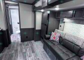 Interior of a modern RV living area with gray walls, dark wood cabinets, and a black leather sofa near a bathroom door on the left