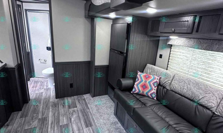 Interior of a modern RV living area with gray walls, dark wood cabinets, and a black leather sofa near a bathroom door on the left