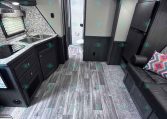 Interior view of a dark-wood RV kitchen: double sink, granite countertop, stove, and cabinets; hallway to bathroom at rear; living area with dark sofa and colorful pillow.
