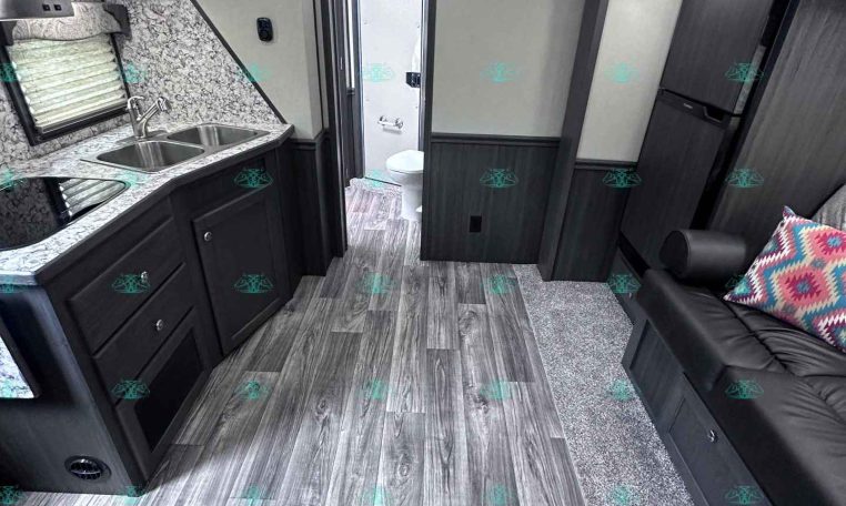Interior view of a dark-wood RV kitchen: double sink, granite countertop, stove, and cabinets; hallway to bathroom at rear; living area with dark sofa and colorful pillow.