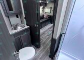 Inside an RV: compact bathroom with granite counter, stainless sink, dark cabinetry, and a nearby toilet, opening to a lounge with a sofa and colorful cushion.
