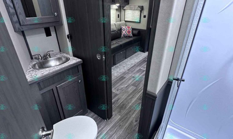 Inside an RV: compact bathroom with granite counter, stainless sink, dark cabinetry, and a nearby toilet, opening to a lounge with a sofa and colorful cushion.