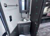 Compact RV bathroom: dark wood vanity with marble countertop, stainless sink, wall mirror, towel bar, and a toilet nearby.