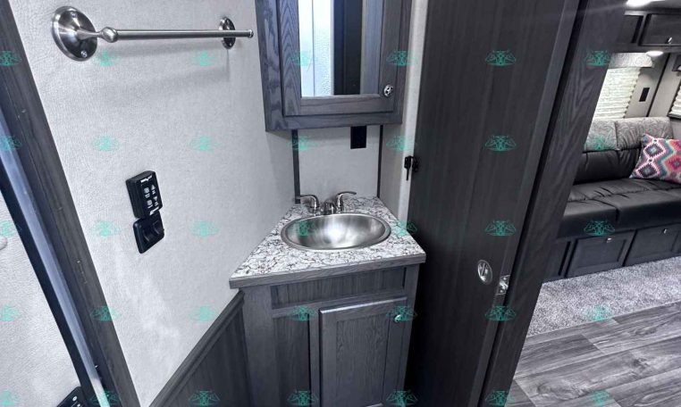 Compact RV bathroom: dark wood vanity with marble countertop, stainless sink, wall mirror, towel bar, and a toilet nearby.