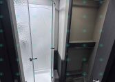 Interior of an RV bathroom with a curved glass shower enclosure on the left and a dark wood cabinet door on the right. Textured white walls and a small round ceiling light visible above.