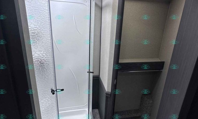 Interior of an RV bathroom with a curved glass shower enclosure on the left and a dark wood cabinet door on the right. Textured white walls and a small round ceiling light visible above.