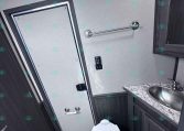 Interior bathroom in a dark-wood trailer: toilet, sink with granite countertop, towel bar, and small door to the left, compact and tiled steps nearby.
