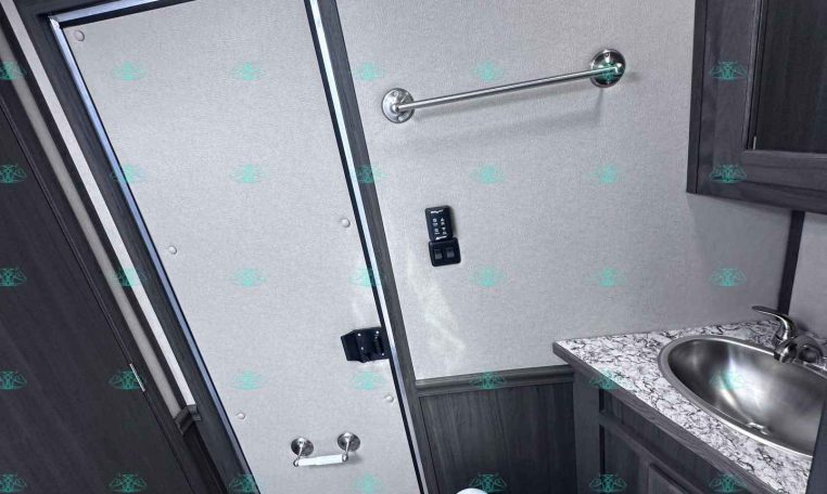 Interior bathroom in a dark-wood trailer: toilet, sink with granite countertop, towel bar, and small door to the left, compact and tiled steps nearby.