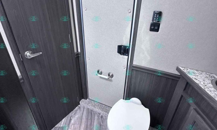 RV bathroom with a white toilet, gray wood-look floor, and dark cabinetry along the walls, plus a towel bar on the right.