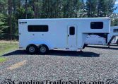 White Shadow Select gooseneck horse trailer with large side window and entry door, parked on gravel with a forest in the background.