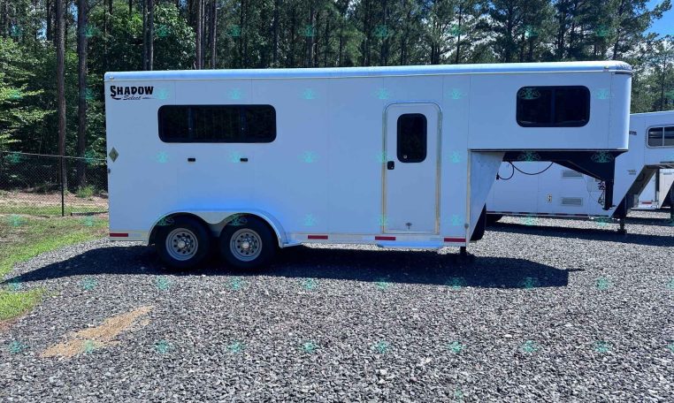 White Shadow Select gooseneck horse trailer with large side window and entry door, parked on gravel with a forest in the background.