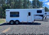 White Shadow Select horse trailer with barred window and entry door on a gravel lot, backed by trees and blue sky.