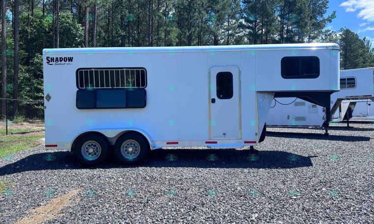 White Shadow Select horse trailer with barred window and entry door on a gravel lot, backed by trees and blue sky.