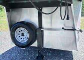 Rear view of a metal utility trailer showing a mounted spare tire, black hoses, and trailer hitch/jack at the front underside area.