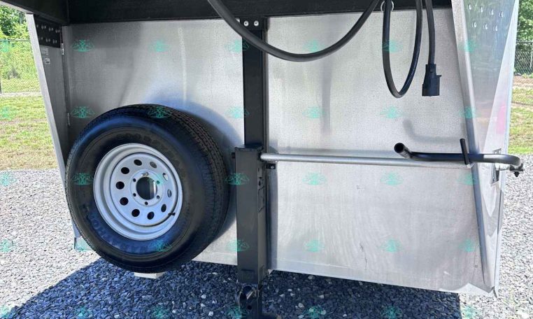 Rear view of a metal utility trailer showing a mounted spare tire, black hoses, and trailer hitch/jack at the front underside area.