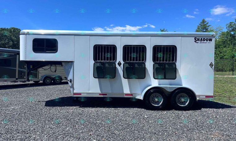 White Shadow Select horse trailer with three barred side windows, parked on a gravel lot under a bright blue sky.