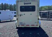 Rear view of a white Shadow Select camper trailer with a large window and branding, parked on gravel with other trailers and trees in the background.