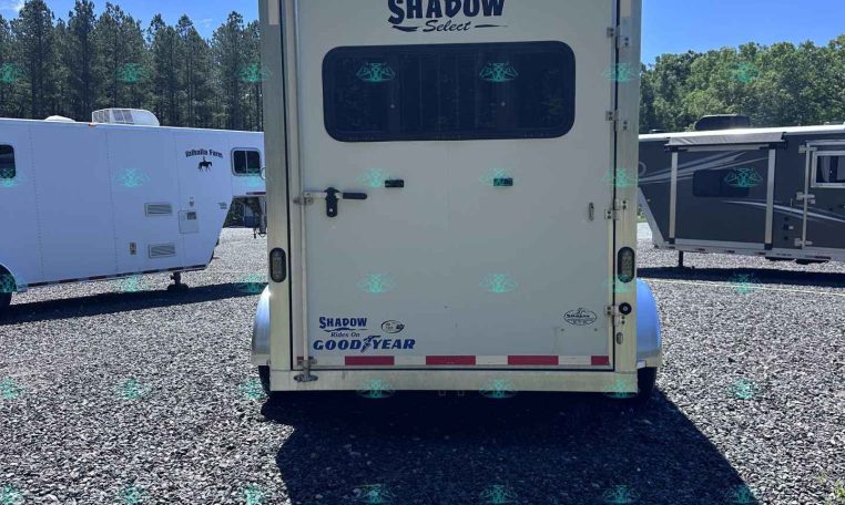 Rear view of a white Shadow Select camper trailer with a large window and branding, parked on gravel with other trailers and trees in the background.