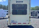 Rear of a white Shadow Select horse trailer with a barred rear window, parked on a gravel lot with other trailers in the background.