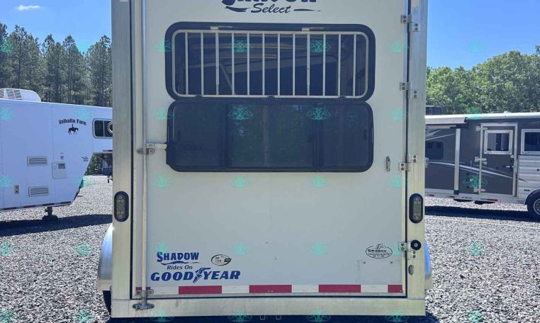 Rear of a white Shadow Select horse trailer with a barred rear window, parked on a gravel lot with other trailers in the background.
