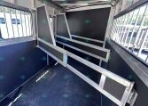 Interior of a metal livestock trailer with partition rails and dark panels, blue non-slip floor, and barred side windows on both sides.