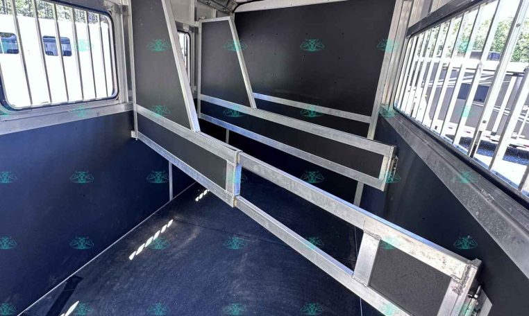 Interior of a metal livestock trailer with partition rails and dark panels, blue non-slip floor, and barred side windows on both sides.