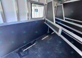Interior of a metal horse trailer with partitioned stalls and a barred window, empty and ready for loading.