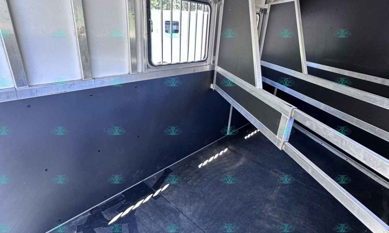 Interior of a metal horse trailer with partitioned stalls and a barred window, empty and ready for loading.
