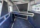 Interior of an empty horse trailer with dark panels, metal frames, and dividers along the sides and ceiling light gaps above.