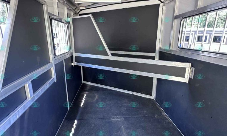 Interior of an empty horse trailer with dark panels, metal frames, and dividers along the sides and ceiling light gaps above.