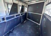 Interior of a black-and-silver horse trailer with divider stalls and windows along the side. Aisle runs down center with metal framing and padded panels on walls.