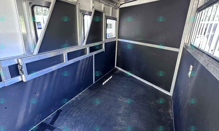 Interior of a black-and-silver horse trailer with divider stalls and windows along the side. Aisle runs down center with metal framing and padded panels on walls.