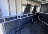Interior of a metal cargo trailer with vertical partitions and small window openings along the side wall, dark floor.