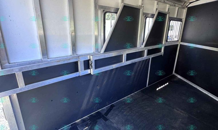 Interior of a metal cargo trailer with vertical partitions and small window openings along the side wall, dark floor.