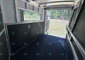 Interior of a metal trailer cabin with dark flooring, storage panels, and a barred window; rear door open to a grassy outdoor area.