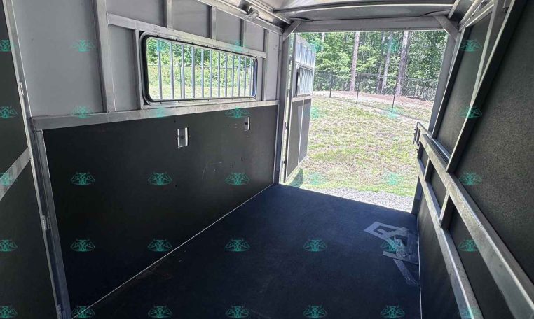 Interior of a metal trailer cabin with dark flooring, storage panels, and a barred window; rear door open to a grassy outdoor area.