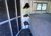 Interior of a metal trailer with black padded wall panels on the left, hanging hooks, and a carpeted raised platform at the right; a window at the far end shows outdoor scenery.