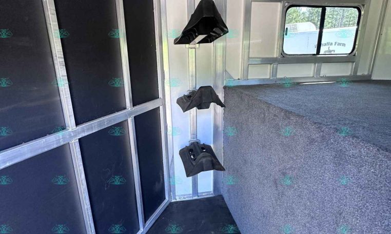 Interior of a metal trailer with black padded wall panels on the left, hanging hooks, and a carpeted raised platform at the right; a window at the far end shows outdoor scenery.